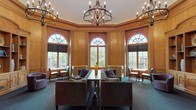 Located at 480 Bedford Road in Chappaqua, New York, this distinguished library space presents a single-floor layout defined by exceptional architectural craftsmanship and a warm, sophisticated ambiance. The interior is characterized by floor-to-ceiling warm-toned wood paneling, intricately detailed with raised moldings, pilasters, and decorative cornices that lend the space a stately, classical character.

Soaring arched windows with fan-light transoms line the perimeter, flooding the room with natural light while offering views of the surrounding greenery. Iron-and-candle-style chandeliers hang from the high ceilings, contributing an air of timeless elegance. A built-in wood bookcase with ornamental crown detailing serves as a central architectural focal point, providing both display and storage.

The main lounge area is furnished with tufted velvet sofas, upholstered armchairs in muted purples and grays, and occasional tables arranged to encourage gathering and conversation. Patterned blue-green carpeting anchors the seating areas throughout. Arched doorways with decorative pediments connect to adjoining corridors, maintaining the cohesive classical aesthetic. The overall atmosphere is refined, quietly formal, and deeply inviting.