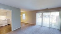 This single-story space features an open, functional layout with distinct living and dining zones flowing seamlessly into one another. The main living area is finished with plush neutral carpeting, while the adjoining room transitions to smooth hardwood-style flooring, providing a clear visual distinction between the two spaces. Walls are painted in soft, neutral tones — a cool grey in the living area and a warm beige in the adjacent room — creating a calm, welcoming ambiance throughout.

Natural light is a defining characteristic of this space. A large sliding glass door fitted with vertical blinds dominates one wall of the living area, offering generous outdoor views and allowing ample sunlight to fill the room. A window in the adjoining room further enhances the brightness, accompanied by a small ceiling-mounted light fixture. HVAC vents are visible in the ceiling, indicating a central climate control system.

The clean, unfurnished state of the space allows for flexible furniture arrangements to suit a variety of lifestyle needs. White baseboards throughout provide a crisp, finished look, complementing the neutral color palette and contributing to the overall tidy, move-in-ready appearance of the space.