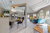 This single-floor communal lounge is designed with a bold, eclectic aesthetic that blends mid-century modern style with coastal-inspired accents. The open-plan layout flows seamlessly between distinct functional zones, all unified by light wood-look flooring and warm greige walls. Soaring vaulted ceilings with exposed white beams define the main lounge area, which is anchored by a teal tufted sofa, sculptural yellow egg chairs, and a live-edge wood coffee table atop a geometric multicolor area rug. A dramatic arched window flanked by large grid-pane windows floods this area with natural light and outdoor views of lush landscaping. A dedicated seating nook features a curved banquette with chevron upholstery surrounding a round concrete table, complemented by woven black rattan chairs and a drum pendant light overhead. The kitchenette area offers a fully equipped service counter with reclaimed wood cabinetry, stainless steel appliances, and a Starbucks coffee machine. Eclectic wall art, including ocean-themed paintings, a decorative fish sculpture, and colorful framed prints, adds personality throughout. Structural columns with wood-accented framing provide subtle architectural definition without interrupting the open, airy flow of the space.