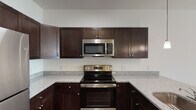 This single-floor unit is designed with a functional and cohesive aesthetic throughout. The kitchen serves as a standout feature, showcasing rich dark-espresso cabinetry with brushed nickel hardware, granite-style speckled countertops, and a full suite of stainless steel appliances including an electric range, over-the-range microwave, and refrigerator. A peninsula with an undermount sink and pendant lighting overhead adds both workspace and visual appeal, while dark hardwood-style flooring grounds the space with warmth and contrast.

The living areas are finished with neutral wall-to-wall carpeting and bright white walls, creating a clean, airy atmosphere. Ceiling fans with integrated lighting are present throughout, contributing to both comfort and functionality. Natural light filters in generously through windows fitted with horizontal blinds.

The unit includes multiple bedrooms, each spacious and well-lit with natural light. The bathrooms offer practical, clean finishes—one features a full soaking tub and shower combination with a pedestal sink, while another includes a standalone walk-in shower with sliding glass doors and a dark-cabineted vanity with matching granite-style countertop. Hardwood-style flooring carries through the bathrooms, maintaining a consistent design language throughout the unit.