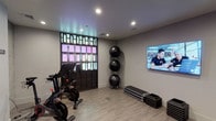 Located at 1601 Broadway in San Diego, this modern fitness and yoga studio offers a thoughtfully designed single-floor space built to support a wide range of workout disciplines. The studio features light wood-style flooring throughout, smooth gray walls, and recessed lighting that creates a bright, clean atmosphere conducive to focused training sessions.

The main workout area is equipped with cycling bikes featuring integrated digital screens, stacked exercise balls, aerobic step platforms, and a wall-mounted flat-screen display for guided workouts. A dedicated strength training corner includes a tiered kettlebell rack and a balance disc, catering to both functional fitness and strength-based training.

Sleek black-framed glass partitions and doors divide the space, lending an industrial-modern aesthetic while maintaining an open, airy feel. A striking backlit panel wall adds a pop of color and visual interest to the interior. The studio also includes a bench seating area and what appears to be an additional adjacent training room, accessible through the glass-paneled entryway.

The space is designed with practicality in mind, featuring smart climate controls, in-ceiling speakers, and clearly posted occupancy guidelines. Overall, the studio combines a polished, contemporary design with a comprehensive array of fitness amenities, making it a versatile environment for yoga, cardio, and strength training.