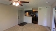 This single-story space in Irving, Texas, offers a practical and functional layout designed for comfortable everyday living. The main living area features wall-to-wall carpeting that adds warmth underfoot, while a ceiling fan overhead provides both airflow and ambient lighting. A textured ceiling runs throughout, adding subtle visual character to the interior.

The living area flows openly toward the kitchen, which is partially separated by a peninsula counter that also functions as a breakfast bar. The kitchen is outfitted with dark espresso-toned cabinetry paired with a mosaic tile backsplash, providing a contemporary contrast to the lighter walls. Black appliances, including a refrigerator, microwave, and range, complement the overall color scheme. Dark flooring in the kitchen ties in with the cabinetry for a cohesive look.

The entry door opens directly into the living space, creating a straightforward and accessible floor plan. A wall-mounted HVAC vent ensures climate control throughout. The neutral wall tones and open sightlines between the living and kitchen areas contribute to a functional, easy-to-navigate environment suited for daily use.