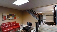 This well-appointed space makes a strong first impression with its refined design and carefully chosen finishes. The centerpiece of the main area is a striking staircase framed by a bold black metal railing with classic newel posts, adding a sense of elegance and structure to the interior. The flooring is a standout feature, showcasing a geometric marble-style pattern in black and white that draws the eye and anchors the space with a sophisticated aesthetic.

The lounge area is furnished with a vibrant red sofa and a sleek black leather accent chair, complemented by a glass-top side table — a combination that balances bold color with contemporary styling. A decorative area rug adds warmth underfoot, while abstract wall art in rich tones contributes a creative touch to the warm, tan-toned walls.

Overhead, large recessed skylight panels flood the space with bright, diffused natural-style light, creating an open and airy atmosphere despite the dropped ceiling grid. Additional recessed lighting continues further into the space, ensuring consistent illumination throughout. The layout feels spacious and flows naturally, with a corridor visible beyond the staircase suggesting further rooms and areas to explore. Overall, the space conveys a professional yet inviting ambiance, suitable for both reception and social functions.