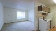 This single-story space features an open-concept layout that connects the living area and kitchen in a practical, functional arrangement. The living area is finished with wall-to-wall carpet in a neutral tone, contributing to a calm and understated ambiance. A large window fitted with vertical blinds allows natural light to fill the room, creating a bright and airy atmosphere. White walls and white-painted baseboards keep the space feeling clean and cohesive throughout.

Adjacent to the living area, the kitchen is partially separated by a half-wall partition, maintaining an open feel while defining the cooking space. The kitchen features a white electric range and is topped with dark wood-toned cabinetry paired with an overhead range hood, providing a subtle contrast to the lighter surroundings. The kitchen flooring transitions to a hard surface, distinguishing it from the carpeted living area.

The textured popcorn ceiling is consistent throughout, adding a classic architectural detail typical of its era. A door located in the living area provides additional access or storage. Overall, the space presents a straightforward, livable layout with practical finishes and adequate natural light.