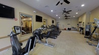 This fitness center offers a comprehensive and thoughtfully arranged workout environment designed to accommodate a variety of exercise routines. The space is outfitted with a row of treadmills, elliptical machines, and stationary cycling equipment, providing ample options for cardiovascular training. Strength and resistance machines are positioned toward the back of the room, rounding out the full range of fitness offerings. The light-toned flooring runs throughout, lending the space a clean and open feel. Recessed lighting is complemented by multiple ceiling fans, ensuring the area remains bright and well-ventilated during workouts. Wall-mounted televisions are strategically placed to keep users entertained while exercising. A large mirror on one wall visually expands the space and allows for form checks during workouts. A window or pass-through opening connects the fitness area to an adjacent lounge or common room, enhancing the sense of openness and community. The pale yellow walls and neutral tones throughout create a welcoming, energizing atmosphere. The overall layout is spacious and practical, with equipment arranged to allow for comfortable movement and safe use of all machines.