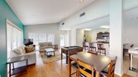 Located at 715 Marie Antoinette St. in Lafayette, Louisiana, this single-floor open-plan space seamlessly integrates a living room, dining area, and kitchen into one cohesive layout. Soaring vaulted ceilings amplify the sense of space throughout, while an abundance of large windows dressed with white horizontal blinds floods the interior with natural light.

The living area is a standout, anchored by a bold teal accent wall that adds a vibrant pop of color against the otherwise neutral palette. Warm hardwood flooring extends through the living and dining zones, complementing the relaxed, welcoming atmosphere. A sliding glass door provides direct access to an outdoor patio area.

The dining area transitions naturally from the living space and features light tile flooring, with room for multiple seating arrangements. A foosball table serves as a recreational centerpiece, lending a social, communal character to the space.

The kitchen is well-appointed with rich dark-stained cabinetry, black granite countertops, stainless steel appliances, and a generous wraparound layout. A raised breakfast bar with bar-height seating connects the kitchen to the dining area, enhancing the open, interactive flow of the space. Recessed lighting throughout ensures consistent illumination, making this a functional and inviting environment overall.