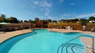 Located at 2100 McAndrew Dr in Raleigh, NC, this outdoor amenity space centers around a generously sized freeform swimming pool with crystal-clear blue water. The pool area is thoughtfully designed for both relaxation and recreation, featuring a wide concrete sun deck lined with multiple lounge chairs ideal for sunbathing. A dedicated clubhouse building with a classic gabled roof, white trim, and French doors anchors one end of the pool area, providing a sheltered gathering spot with outdoor seating. The space is enclosed by a sturdy black iron fence, ensuring safety and a clean aesthetic that complements the surrounding landscaping. Lush green lawns, mature trees, and manicured shrubbery frame the area, creating a serene, park-like atmosphere. Comfortable outdoor seating options, including cushioned lounge sofas with decorative pillows and umbrella shade, are thoughtfully positioned around the pool deck. Safety equipment, including a life ring station, is conveniently mounted along the fence. The backdrop reveals a rolling residential hillside dotted with homes and mature trees, adding a picturesque, community-oriented setting to this well-appointed outdoor retreat. The space enjoys abundant natural light under open skies, making it an inviting destination throughout the day.