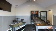 This single-floor game room is purpose-built for recreation and entertainment, offering a well-appointed space that invites friendly competition. The room's most striking architectural feature is its vaulted ceiling, which adds a sense of openness and volume, complemented by a ceiling fan that keeps the space comfortable during lively play sessions. A two-tone wall treatment — with a painted upper half and a gray shiplap-style lower wainscoting — gives the room a polished, stylish character.

The space is outfitted with a classic foosball table, perfect for head-to-head matchups, and a full arcade-style basketball shootout game complete with multiple balls and a backboard display, making it a standout centerpiece. A pair of adjustable bar-height stools provide seating for spectators or those waiting their turn. The dark carpeted flooring anchors the room and adds a cozy contrast to the lighter walls. HVAC vents are neatly integrated into the ceiling, ensuring year-round comfort. Whether used for casual gatherings or competitive game nights, this dedicated game room delivers an engaging and energetic atmosphere for all ages.