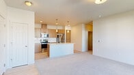 This single-floor residential unit showcases a contemporary design with a clean, neutral color palette throughout. The open-concept kitchen and living area serve as the heart of the home, where light wood cabinetry, stainless steel appliances — including a refrigerator, range, and built-in microwave — and a crisp white tile backsplash create a modern and functional cooking space. A central island with a light quartz countertop and an integrated sink adds both prep space and a natural gathering point. Pendant lighting above the island and recessed ceiling lights provide warm, layered illumination. Light hardwood-style flooring in the kitchen transitions seamlessly into plush carpeting in the living and bedroom areas. The primary bedroom is generously proportioned, featuring sliding glass door access to an outdoor balcony with exterior views, along with a large window that fills the room with natural light. A spacious walk-in closet is outfitted with multi-level wire shelving and hanging rods, offering ample storage. The bathroom features light wood cabinetry, a white countertop vanity, a large mirror, and a full tub-shower combination with clean white surround. An additional bedroom provides comfortable, carpeted space with easy closet access. Overall, the unit presents a bright, well-organized living environment with modern finishes throughout.