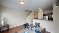 This two-storey space offers a well-organized, open-concept layout that connects the main living areas seamlessly. The dining area is positioned centrally on the main floor, accommodating a full-sized dining table with seating for multiple guests. Hardwood-style flooring runs throughout the main level, adding warmth and continuity to the space.

Adjacent to the dining area, the kitchen features a raised breakfast bar with a stone or granite-style countertop, dark cabinetry, and stainless steel appliances, including a refrigerator. Recessed and flush-mounted ceiling lighting illuminate both the kitchen and dining zones effectively.

A staircase with a white railing rises prominently from the main floor, providing access to the upper level while also serving as a visual dividing element between the living and kitchen areas. The neutral beige wall tones throughout contribute to a clean, understated aesthetic. The overall layout prioritizes functionality and flow, making the main floor well-suited for both everyday living and casual entertaining.