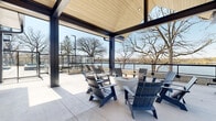 This contemporary single-level outdoor amenity space is defined by its striking modern architecture and exceptional waterfront setting. The centerpiece is a generously sized covered terrace featuring bold black steel framing and a warm wood-paneled ceiling, creating a sheltered yet open-air gathering area. Navy Adirondack chairs are arranged around a round fire pit, offering a comfortable and inviting atmosphere with panoramic views of the adjacent lake and mature oak trees beyond the glass railing perimeter.

Extending from the covered terrace, an open-air patio provides additional lounge seating with neutral-toned sofas and chairs, ideal for taking in unobstructed waterfront views. Large floor-to-ceiling glass walls connect the covered outdoor structure to interior spaces, allowing natural light to flow freely while maintaining visual continuity between indoor and outdoor areas.

A well-appointed swimming pool occupies a dedicated section of the terrace, lined with lounge chairs and framed by the waterfront backdrop. A built-in gas grill is also available for outdoor entertaining. Concrete pavers throughout unify the various zones, while the clean, minimalist design aesthetic and proximity to the water lend the space a calm, resort-like ambiance.