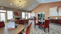 This single-floor community lounge is designed to foster a warm and inviting atmosphere, combining functional layout with thoughtful décor. The space is anchored by soaring vaulted ceilings that lend an open, airy feel, enhanced by a striking geometric chandelier with exposed Edison-style bulbs that serves as a central focal point. Recessed lighting complements the natural light filtering through multiple windows, keeping the space bright and welcoming.

A neutral-toned carpet with a subtle floral pattern runs throughout, grounding the room's warm color palette. An accent wall painted in a rich terracotta hue adds depth and visual interest, paired with curated artwork that reinforces the contemporary aesthetic. A classic fireplace with a white mantel anchors one end of the space, creating a cozy seating area furnished with plush armchairs in earthy tones.

Dining-height tables surrounded by tufted red leather chairs are positioned throughout, offering flexible seating arrangements. A built-in niche along one wall features teal-painted shelving, providing display and storage space. A small beverage station adds convenience, making this lounge well-suited for casual gatherings and community events alike.