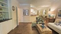 This single-floor residence is designed with an open-concept layout that seamlessly connects the living, dining, and kitchen areas into one cohesive space. Rich, dark hardwood-style flooring runs throughout, providing a warm and grounding foundation that contrasts elegantly with the light, neutral-toned walls. Crown molding adds a refined architectural detail, lending a polished character to each room.

The living area is anchored by a rustic pallet-style coffee table and a boldly patterned accent chair with a coordinating ottoman, offering a relaxed yet stylish seating arrangement. Adjacent to the living space, a pass-through counter connects to the kitchen, which features white cabinetry and hints of a contrasting countertop. The kitchen's layout supports easy interaction with the surrounding living and dining areas.

The dining area is illuminated by a pendant chandelier, creating an inviting atmosphere for meals. Framed artwork and decorative wall accents contribute to the overall aesthetic. A hallway near the entry leads to additional rooms, with white panel doors maintaining a clean, consistent look throughout. The space as a whole feels bright, functional, and thoughtfully appointed.