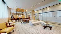 This upscale amenity lounge is designed with a refined, contemporary aesthetic that seamlessly blends multiple social and recreational zones within a single open-plan floor. The space is anchored by a herringbone-patterned light hardwood floor that extends throughout, contrasting elegantly with warm wood-paneled walls and a recessed wood-clad ceiling detail in the billiards area.

The games zone features a sleek white billiards table positioned beneath a sculptural linear pendant light, with a wall-mounted television and a built-in wet bar with dark wood cabinetry nearby. A glazed partition wall with floor-to-ceiling black-framed glass panels creates a transparent boundary, maintaining visual openness while defining separate areas.

The lounge zone is furnished with a large sectional sofa in neutral tones, mid-century-inspired wooden dining chairs around a round table, and mustard-yellow accent armchairs arranged on a patterned area rug. A full media wall with open white shelving and a wall-mounted television anchors this seating area. Sheer floor-to-ceiling curtains diffuse natural light along the perimeter, contributing to the bright, airy ambiance throughout the space.