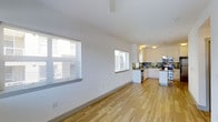 This single-floor space offers an open-concept layout that seamlessly connects the living area to the kitchen. Light hardwood flooring runs throughout, lending warmth and continuity to the interior. The living area features clean white walls, vaulted ceilings that enhance the sense of openness, and two windows fitted with horizontal blinds that allow for adjustable natural light.

The kitchen is well-appointed with white cabinetry, dark stone countertops, and a decorative tile backsplash in earthy green and neutral tones. Stainless steel appliances, including a range and dishwasher, are integrated into the design. The kitchen is arranged in an L-shaped configuration, providing ample counter and storage space. Overhead lighting illuminates the workspace effectively, with pendant and flush-mount fixtures adding a functional touch.

Overall, the space presents a clean, modern aesthetic with a bright and airy ambiance. The open floor plan makes the interior feel spacious and versatile, suitable for a variety of living arrangements. The combination of natural light, quality finishes, and practical kitchen design contributes to a comfortable and functional environment.