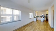 This single-floor space offers an open-concept layout that seamlessly connects the living area to the kitchen. Light hardwood flooring runs throughout, lending warmth and continuity to the interior. The living area features clean white walls, vaulted ceilings that enhance the sense of openness, and two windows fitted with horizontal blinds that allow for adjustable natural light.

The kitchen is well-appointed with white cabinetry, dark stone countertops, and a decorative tile backsplash in earthy green and neutral tones. Stainless steel appliances, including a range and dishwasher, are integrated into the design. The kitchen is arranged in an L-shaped configuration, providing ample counter and storage space. Overhead lighting illuminates the workspace effectively, with pendant and flush-mount fixtures adding a functional touch.

Overall, the space presents a clean, modern aesthetic with a bright and airy ambiance. The open floor plan makes the interior feel spacious and versatile, suitable for a variety of living arrangements. The combination of natural light, quality finishes, and practical kitchen design contributes to a comfortable and functional environment.