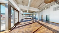 This single-floor commercial space combines industrial and contemporary design elements to create a bright, versatile environment. The main open area is defined by striking wide-plank hardwood flooring in rich, warm tones that contrast beautifully with the exposed concrete ceiling above. Large-diameter HVAC ductwork runs overhead alongside track lighting, lending an industrial-chic character to the space. Floor-to-ceiling storefront windows and glass entry doors line the street-facing façade, drawing in abundant natural light and offering clear views of the surrounding neighborhood streetscape. Concrete structural columns punctuate the open floor plan, reinforcing the raw architectural aesthetic while maintaining a sense of spaciousness throughout. A secondary open room at the rear features white-painted walls and its own distinctive pendant lighting fixtures, including an ornate globe chandelier and delicate hanging bird accents, creating an inviting and adaptable space. The facility also includes a clean, ADA-accessible restroom finished with large-format tile flooring, grab bars, and a skylight that provides additional natural illumination. Overall, the space presents a polished yet industrial ambiance well-suited for retail, studio, gallery, or creative office use.