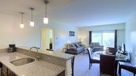 This single-floor residence is designed with an open-concept layout that integrates the kitchen, dining, and living areas into one cohesive, flowing space. The kitchen is anchored by a granite countertop peninsula with an undermount sink, providing both functional workspace and a natural dividing point between the cooking and living zones. Pendant lights hang above the peninsula, adding a warm, focused glow to the area. The dining space sits adjacent to the kitchen, furnished with a round table and dark upholstered chairs, creating an intimate setting for meals. The living area extends toward a large window dressed with curtains, allowing generous natural light to fill the room and offering views of surrounding greenery. Plush carpeting throughout the living space adds warmth and comfort underfoot. Neutral-toned walls and light-colored ceilings contribute to an airy, open ambiance. A doorway visible from the main living area leads to additional rooms beyond, suggesting a practical and well-organized floor plan. Overall, the space presents a clean, modern aesthetic with a comfortable and welcoming atmosphere.