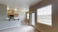 This single-floor space showcases an open-concept layout that seamlessly connects the living area and kitchen. The living area is finished with neutral-toned carpet, while the kitchen transition is marked by light hardwood-style flooring. Warm beige walls complement the overall palette, creating a cohesive and inviting atmosphere throughout.

The kitchen features medium-toned wood cabinetry, a built-in microwave, and a range. A peninsula counter with bar-height seating extends into the living space, making it ideal for casual dining or entertaining. A pendant light fixture hangs above the peninsula, adding a subtle design accent.

The living area is bathed in natural light, courtesy of multiple large windows fitted with horizontal blinds. A sliding glass door provides direct access to an outdoor balcony, offering views of the surrounding area and enhancing the sense of openness. A flush-mount ceiling light provides additional illumination. Clean white baseboards and a neutral ceiling complete the well-maintained, move-in-ready aesthetic of this functional and light-filled space.