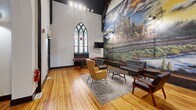 Located at 938 E Fourth St in Bethlehem, PA, this thoughtfully converted historic church blends architectural heritage with modern communal living. The open main floor retains the grandeur of its original design, highlighted by soaring vaulted ceilings with dark exposed wooden beams and a striking dark-trimmed Gothic arch that serves as a dramatic interior passageway. Original stained glass windows cast colorful light into the space, preserving the building's ecclesiastical character while complementing its contemporary use.

The centerpiece of the main gathering area is a sweeping floor-to-ceiling mural depicting Bethlehem's iconic steel industry alongside a vivid landscape, serving as both a tribute to local history and a stunning visual anchor. Warm-toned hardwood floors run throughout, lending continuity and warmth to the space. Seating areas are thoughtfully arranged with mid-century modern lounge chairs, sofas, and coffee tables, while dining tables and a shuffleboard table offer additional recreation and gathering options.

A kitchen provides functional support to the space, and a staircase connects to additional levels. Wall-mounted accent lighting complements the natural light filtering through the stained glass. The overall ambiance is one of creative, community-oriented design — honoring the building's storied past while delivering a vibrant, welcoming environment.