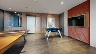 Located at 560 Rideau St in Ottawa, this contemporary games and lounge room offers an elevated social experience in a compact yet thoughtfully designed space. The room is finished with warm hardwood-style flooring throughout, complemented by a sophisticated palette of deep slate-blue and charcoal walls accented by a striking red diamond-patterned upholstered feature wall that anchors the entertainment area. A wall-mounted flat-screen television sits within this bold accent panel, creating a focused media and recreation zone. The space is equipped with a professional foosball table and a full-sized ping pong table, providing ample entertainment options for residents and guests. A cozy dining nook furnished with a marble-top table and plush upholstered chairs offers a comfortable spot to gather, framed by floor-to-ceiling windows that flood the room with natural light and showcase panoramic views of the Ottawa skyline. Warm wood paneling along one wall adds texture and warmth, while recessed ceiling lighting ensures a clean, modern finish. Sheer curtains soften the generous window line, enhancing the relaxed yet refined ambiance. This well-appointed amenity space combines recreational functionality with polished interior design, making it an inviting destination within the building.