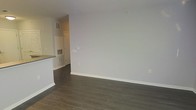 This single-floor space presents a clean, modern interior defined by a cohesive neutral palette and quality finishes. The main living area is finished with rich, dark hardwood-style flooring that contrasts elegantly with the light grey walls and crisp white baseboards, creating a polished, contemporary aesthetic. The open layout allows for flexible furniture arrangements, and the uncluttered design gives the space a sense of openness and ease.

Adjacent to the living area, a partial view of the kitchen reveals a countertop overhang that functions as a convenient peninsula or breakfast bar, subtly separating the kitchen from the living space while maintaining an open feel. The kitchen area features consistent flooring and cabinetry that complements the overall design language of the unit.

Practical elements are thoughtfully integrated throughout, including ceiling vents for climate control and well-placed electrical outlets along the walls. A hallway visible from the main living space suggests additional rooms or utility areas beyond, contributing to a functional and well-organized floor plan. The overall ambiance is fresh, neutral, and move-in ready.