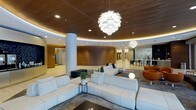 This single-floor space showcases a thoughtfully designed open-concept lobby and lounge area that blends modern architecture with warm, inviting materials. The ceiling is a standout feature, clad in rich wood paneling with a curved, recessed tray design that incorporates subtle ambient lighting along its edges, creating a soft, welcoming glow throughout the space.

Dramatic pendant light fixtures serve as focal points overhead, featuring layered, sculptural designs that draw the eye upward and add an artistic dimension to the interior. Recessed lighting complements these statement pieces, ensuring the space is evenly and elegantly illuminated.

The lounge area is anchored by expansive white sectional sofas arranged on a large area rug, offering generous and comfortable seating. Orange accent chairs introduce a bold pop of color, balancing the otherwise neutral palette. Cylindrical white columns punctuate the open floor plan, contributing to the architectural rhythm of the space.

Warm walnut wood wall panels line the curved central core, reinforcing the cohesive design language. In the background, a bright, glass-enclosed workspace or meeting area is visible, highlighting the space's functional versatility alongside its refined aesthetic.