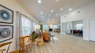 Located at 174 Emery Ave NW in Concord, NC, this single-story space presents a well-designed, open-concept layout finished with light-toned luxury vinyl plank flooring and crisp white walls throughout. The main living area is generously proportioned, featuring vaulted ceilings with recessed lighting and an abundance of natural light delivered through multiple large windows dressed with linen curtains and plantation-style blinds. The living space comfortably accommodates a full seating arrangement and flows seamlessly into the dining and kitchen areas, creating a cohesive and functional environment.

The kitchen is outfitted with white shaker-style cabinetry, granite countertops, stainless steel appliances, and a substantial island with seating — ideal for casual dining or collaborative use. Vaulted ceilings continue into this area, reinforcing the open, airy character of the space.

A dedicated office or meeting room is separated from the main area by glass sliding doors, offering privacy while maintaining visual connectivity. This room features soaring ceilings, a modern globe chandelier, a built-in desk, and gallery-style wall art, creating a polished and professional atmosphere well-suited for focused work or client meetings.