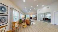 Located at 174 Emery Ave NW in Concord, NC, this single-story space presents a well-designed, open-concept layout finished with light-toned luxury vinyl plank flooring and crisp white walls throughout. The main living area is generously proportioned, featuring vaulted ceilings with recessed lighting and an abundance of natural light delivered through multiple large windows dressed with linen curtains and plantation-style blinds. The living space comfortably accommodates a full seating arrangement and flows seamlessly into the dining and kitchen areas, creating a cohesive and functional environment.

The kitchen is outfitted with white shaker-style cabinetry, granite countertops, stainless steel appliances, and a substantial island with seating — ideal for casual dining or collaborative use. Vaulted ceilings continue into this area, reinforcing the open, airy character of the space.

A dedicated office or meeting room is separated from the main area by glass sliding doors, offering privacy while maintaining visual connectivity. This room features soaring ceilings, a modern globe chandelier, a built-in desk, and gallery-style wall art, creating a polished and professional atmosphere well-suited for focused work or client meetings.