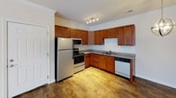 This single-floor residence offers a practical and cohesive layout, with distinct living, sleeping, and functional zones that flow naturally from one to the next. The kitchen is a standout feature, outfitted with rich cherry wood cabinetry, granite-style countertops, and a full suite of stainless steel appliances including a refrigerator, electric range, over-the-range microwave, and dishwasher. Wood-look flooring grounds the kitchen area, which is illuminated by track lighting and crown molding details that add a polished finish.

The living area transitions seamlessly from the kitchen, featuring soft wall-to-wall carpeting and neutral tones that create a warm, inviting atmosphere. A pendant light fixture adds a subtle decorative touch near the kitchen boundary.

The bedroom is generously sized with plush carpeting, a window fitted with blinds for natural light control, and access to a walk-in closet. French doors open from the bedroom, providing additional light and outdoor access.

The full bathroom includes a soaking tub with shower, a chrome towel bar, a toilet, and a vanity with a rich wood cabinet base, white countertop, and a well-lit mirror above — completing this functional and comfortable residence.