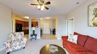 This single-story residence features an open-concept layout that seamlessly connects the living room, dining area, and kitchen, creating a spacious and functional flow throughout. The living area is finished with plush carpeting and is anchored by a ceiling fan with integrated lighting, providing both comfort and airflow. Neutral gray walls complement the warm tones of the furnishings and create a versatile backdrop throughout the space.

The kitchen, visible from the living room, features dark wood cabinetry, a black refrigerator, and a raised breakfast bar with seating, making it ideal for casual dining. Tile flooring runs through the kitchen and transitional areas, offering a practical contrast to the carpeted living space. Structural columns subtly define the boundary between the living and kitchen zones while maintaining the open feel.

A dining area sits adjacent to the kitchen, accommodating a small table and chairs. Two interior doors lead to additional rooms, suggesting private spaces such as bedrooms or a bathroom. The overall ambiance is warm and inviting, with an efficient, well-organized layout suited for everyday living.