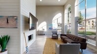 This single-floor space showcases a thoughtfully designed coastal aesthetic, combining architectural grandeur with relaxed, inviting interiors. The main lounge area immediately captures attention with its dramatically tall ceilings and a series of tall arched windows that flood the space with abundant natural light, casting sweeping arcs of sunlight across the light-toned hardwood-style flooring. A striking shiplap accent wall anchors one side of the room, featuring a sleek wall-mounted flat-screen television above a minimalist fireplace. Coastal-themed artwork, including vivid surf and vintage car paintings, reinforces the beach-inspired design throughout.

Comfortable seating arrangements, including a tufted sofa, accent chairs, and a natural jute area rug, create a welcoming lounge environment. A decorative oar leans against the fireplace wall, adding a nautical touch. An upper mezzanine level with black iron railing is visible overhead, adding vertical dimension to the space.

Beyond the main lounge, the layout opens into additional multipurpose areas featuring recessed lighting, white structural columns, and glazed interior doors leading to auxiliary rooms. A casual high-top table and seating area, an ornate sideboard, and tasteful greenery are distributed throughout, contributing to the space's cohesive, airy coastal character.