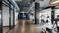 Located at 679 S Hobart Blvd in Los Angeles, The Pearl is a expansive, full-service fitness facility designed with a modern industrial aesthetic. The single-floor space encompasses a generous footprint, divided into distinct functional zones that cater to a wide range of workout preferences.

The main gym floor features large-format tile flooring and an impressive array of strength and cardio equipment, including treadmills, ellipticals, cable machines, and a power rack. Exposed ductwork and dark ceiling infrastructure lend an industrial character, while oversized drum pendant lights provide warm, focused illumination. Floor-to-ceiling windows along the perimeter flood the space with natural light and offer outdoor views.

A dedicated cycling studio houses rows of spin bikes arranged before full-wall mirrors, with a ballet barre running the length of the room — suitable for both cycling and barre-style classes. Adjacent to it, a spacious group fitness and dance studio features wood-look flooring, mirrored walls, and a wall-mounted display screen, offering a clean, versatile environment for classes or open movement.

A functional training area includes a heavy bag, TRX suspension trainers, medicine balls, and a bench press zone. The entry area features a Brita hydration station and a branded welcome mat, reinforcing the facility's polished, amenity-forward identity throughout.