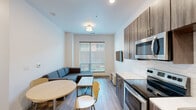 This contemporary single-floor unit is designed with a clean, modern aesthetic and a functional, open layout. The kitchen is a standout feature, equipped with a full suite of stainless steel appliances including an electric range, over-the-range microwave, dishwasher, and refrigerator. Dark wood-grain cabinetry with brushed metal hardware extends to the ceiling, complemented by white quartz countertops and a stylish hexagonal tile backsplash. Light wood-look flooring runs throughout, lending warmth and continuity to the space. The kitchen flows seamlessly into the dining and living areas, where a round dining table and a sectional sofa anchor the room. A large window floods the living space with natural light and provides views of the surrounding neighborhood. The bedroom is well-appointed with oversized windows on two walls, offering abundant daylight and exterior views. A built-in desk and shelving unit make it a practical space for work or study. The bathroom maintains the unit's cohesive design with matching wood-grain cabinetry, white quartz surfaces, light wood-look flooring, and a grab bar for added accessibility. Throughout the unit, white walls and flush-mount lighting contribute to a bright, airy atmosphere.