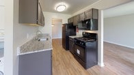 This single-floor residence offers a practical and comfortable layout designed for everyday living. The kitchen is a standout feature, outfitted with dark espresso cabinetry, granite-look countertops, and a full suite of black appliances including a gas range, over-the-range microwave, and refrigerator. Wood-look flooring runs throughout the kitchen, adding warmth and easy maintenance to the space.

The living and dining areas are open and generously sized, with the same wood-look flooring continuing seamlessly from the kitchen. A pendant light fixture defines the dining area, while neutral gray walls and white trim create a clean, cohesive aesthetic throughout.

The bedroom is a comfortable retreat, featuring plush wall-to-wall carpeting, a large window that brings in natural light and outdoor views, and a bifold-door closet offering ample storage space. The neutral tones carry through here as well, lending a calm and restful atmosphere.

The bathroom is finished with white square tile surrounding a full-size soaking tub and shower combination, complete with chrome fixtures. The bright, clean design keeps the space feeling fresh and functional.

Overall, this residence presents a straightforward and well-maintained interior with consistent finishes, practical amenities, and a layout that balances open communal spaces with private living areas.