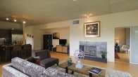 This single-floor space showcases a thoughtfully designed interior with a contemporary yet welcoming ambiance. The open-concept living area serves as the heart of the space, anchored by a striking fireplace encased in a sleek tile surround, which adds both warmth and a refined focal point to the room. Warm hardwood flooring flows throughout, complementing the neutral-toned walls and creating a cohesive, inviting atmosphere.

The living area is generously proportioned, offering ample room for comfortable seating arrangements. A plush area rug grounds the space, while a rustic wooden coffee table and decorative sculptural accents contribute to the eclectic, curated aesthetic. Framed artwork displayed above the fireplace and along the walls adds a personal and artistic touch to the environment.

The ceiling features a distinctive paneled design, lending an architectural character that distinguishes the space. Recessed ventilation and subtle overhead lighting ensure both comfort and functionality. A media console with storage is neatly integrated into the layout, providing practical organization without disrupting the visual flow. The open sightlines extend across the room, reinforcing a sense of spaciousness and ease of movement throughout this well-appointed single-level space.