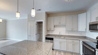 Located at 6701 Fox Rd in Raleigh, NC, this single-floor residence offers a thoughtfully designed living space with modern, contemporary finishes throughout. The kitchen serves as a central highlight, showcasing light granite countertops with subtle speckled detailing that extend across both the main counter surfaces and a generous island. Soft gray shaker-style cabinetry provides ample storage, complemented by sleek brushed nickel hardware for a clean, cohesive aesthetic. A stainless steel dishwasher and a single-basin undermount sink with a modern faucet complete the functional kitchen setup. Pendant lighting with cylindrical frosted glass shades hang above the island, casting warm, focused light over the workspace. The flooring features wood-look planks in a light, neutral tone, adding warmth and continuity throughout the space. A walk-in pantry with built-in wire shelving offers excellent organizational capacity and is conveniently accessible directly from the kitchen. The walls are finished in a soft, neutral beige tone, creating a bright and welcoming ambiance that complements the overall open-concept layout. This residence combines practicality with modern style, making it an inviting and well-appointed home.