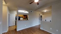 This single-floor space showcases an open-concept layout designed to maximize connectivity between living and kitchen areas. The flooring throughout is a warm hardwood-style material that adds a cohesive and inviting feel to the space.

The kitchen is partially separated from the main living area by a half-wall featuring a distinctive arched pass-through window, which allows natural sightlines between both spaces while maintaining a sense of definition. The kitchen is equipped with medium-toned wood cabinetry, dark appliances including a refrigerator and microwave, and a countertop that extends to the pass-through, offering casual seating or serving opportunities.

The living area is spacious and open, anchored by a ceiling fan with integrated lighting that provides both comfort and illumination. Neutral-toned walls throughout keep the space feeling bright and versatile.

A built-in shelving unit with arched detailing is visible along one wall, adding both storage and architectural character. A half-wall with decorative trim further divides areas of the space while maintaining an airy, open feel. Additional doorways suggest access to private rooms beyond the main living space.