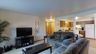 This single-floor residence offers a practical and comfortable layout with an open flow between the main living area and the kitchen. The living room is carpeted and features neutral wall tones with a flush ceiling light, creating a warm, understated ambiance. The space accommodates a plush sectional-style sofa arrangement centered around a rectangular coffee table and a media console supporting a flat-screen television. A small gallery wall with framed artwork adds a personal touch to the room.

A wide opening connects the living room directly to the kitchen and dining area, enhancing the sense of openness. The kitchen is equipped with warm-toned wooden cabinetry, overhead lighting, and a countertop workspace. Appliances are visible along the back wall, and a small dining table with chairs is positioned near the kitchen, making it a convenient space for casual meals.

A wood-paneled door visible in the background provides access to additional rooms or storage. Overall, the space maintains a cohesive, neutral palette throughout, emphasizing functionality and ease of everyday living within a compact, well-organized single-floor layout.