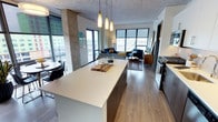 This contemporary urban residence showcases an industrial-modern aesthetic defined by exposed concrete ceilings throughout, warm hardwood-style flooring, and expansive floor-to-ceiling windows that frame striking city and skyline views. The open-concept kitchen and living area form the heart of the space, where a large central island with a clean white quartz surface anchors the layout. The kitchen features a two-tone cabinetry design—combining white upper cabinets with dark wood lower units—complemented by stainless steel appliances, a subway tile backsplash, and elegant pendant lighting above the island. The living area flows naturally from the kitchen, bathed in natural light from oversized windows and a glass door leading to a private balcony, offering panoramic urban views. The bedrooms maintain the industrial character with exposed concrete ceilings and large windows. The primary bedroom includes a walk-in closet with built-in shelving. Bathrooms are clean and modern, featuring tile flooring, dark wood vanities with white countertops, and a powder room with a minimalist design. Throughout the residence, exposed concrete structural columns add authentic industrial character, creating a cohesive, contemporary urban aesthetic.