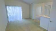 This single-floor space features an open and straightforward layout with a clean, neutral aesthetic throughout. The main living area is finished with wall-to-wall carpet in a light beige tone, contributing to a soft and understated atmosphere. Walls are painted in a crisp white, enhancing the sense of brightness throughout the room.

A large window dressed with vertical blinds allows natural light to filter into the living area, illuminating the space evenly. A ceiling vent is visible, indicating a central HVAC system for climate control.

To the right of the main living area, a partial countertop or breakfast bar element is visible, suggesting an adjacent kitchen or transitional space. A doorway leads further into the unit, offering a glimpse of an additional carpeted room beyond, indicating connected living spaces within the single-floor plan.

The overall design is minimal and functional, with straightforward finishes suited for practical everyday living. The open flow between spaces and the abundance of natural light give the interior a clean, uncluttered feel.