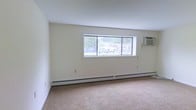 This single-floor unit offers a straightforward and functional layout designed for comfortable everyday living. The main living area is finished with wall-to-wall carpeting, providing a warm and cushioned underfoot feel throughout the space. Neutral cream-toned walls create a clean, bright backdrop that complements the natural light streaming in through a wide, horizontally oriented window. This window serves as a focal point of the room, offering views of the surrounding outdoor environment and allowing ample daylight to fill the interior. Climate control is well-addressed with a continuous baseboard heating system running along the exterior wall, ensuring even warmth distribution during colder months, while a wall-mounted air conditioning unit provides cooling relief in warmer seasons. The smooth white ceiling contributes to the open, airy feel of the room. The overall ambiance is understated and practical, offering a neutral canvas that can easily be personalized to suit a variety of lifestyles. The space is well-suited for those seeking a manageable, easy-to-maintain living environment with essential comforts built in.