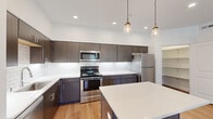 This single-story home offers three bedrooms, two bathrooms, and a well-designed open-concept kitchen and living area finished with consistent light hardwood flooring throughout. The kitchen serves as a central highlight, featuring dark wood cabinetry with brushed nickel hardware, a white quartz countertop, a white subway tile backsplash, and a full suite of stainless steel appliances including a range, microwave, and refrigerator. A center island with pendant lighting above provides additional prep and casual dining space. An open pantry with built-in shelving offers convenient storage directly off the kitchen. The living area is bright and open, equipped with a wall-mounted mini-split HVAC unit and access to natural light through windows with blinds. The primary bedroom is a comfortable size, featuring vaulted ceilings, a ceiling fan, multiple windows with outdoor views, and a walk-in closet with double hanging rods and natural light. The additional bedrooms are clean and simply appointed with hardwood floors and white walls. The bathrooms include a wide dark wood vanity with quartz countertop, a large mirror with overhead lighting, and a walk-in shower. Overall, the space presents a clean, contemporary aesthetic throughout.