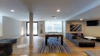 This single-floor amenity space offers a well-appointed and contemporary environment designed for socializing and recreation. At the heart of the room sits a full-size billiards table, positioned atop a bold, multi-toned striped area rug that anchors the space and adds a dynamic visual element to the wide-plank hardwood flooring beneath. A stylish geometric chandelier hangs above the billiards area, complementing the modern aesthetic throughout.

Large windows along the far wall flood the interior with natural light, creating an open and inviting atmosphere. A comfortable lounge area with ample seating is arranged to one side, ideal for relaxed conversation or casual gatherings. An abstract wall-mounted artwork adds a splash of color and artistic character to the neutral-toned walls.

Structural columns are integrated seamlessly into the layout, while recessed lighting supplements the natural illumination. Glass-paneled entry doors provide visual connectivity to adjacent areas or outdoor spaces, further enhancing the sense of openness. The overall design balances clean architectural lines with carefully selected furnishings and décor, resulting in a polished, functional, and welcoming communal space.
