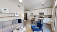 This stylish single-floor residence showcases a thoughtfully designed open-concept layout that seamlessly connects the living and kitchen areas into one cohesive, functional space. The interior is characterized by crisp white walls, high ceilings, and recessed lighting that collectively enhance the sense of openness and brightness throughout.

The kitchen is a standout feature, outfitted with sleek white cabinetry, stainless steel appliances including a refrigerator, microwave, and gas range, and a light-toned tile backsplash that adds subtle texture. A peninsula with seating for two or three serves as both a practical workspace and a casual dining area, anchoring the kitchen within the open floor plan.

The living area is warm and inviting, furnished with a neutral sofa accented by navy blue throw pillows and complemented by a bold blue accent chair that introduces a pop of color. A woven area rug defines the seating zone, while a wall-mounted shelf adds a minimalist decorative touch. Light-toned flooring flows consistently throughout, tying the spaces together. The overall ambiance is modern and polished, balancing clean contemporary design with comfortable, livable details.