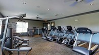 This fitness center offers a comprehensive workout environment designed to accommodate a variety of exercise routines. The space is outfitted with a generous selection of cardio equipment, including a row of treadmills and elliptical machines by TotaLife, positioned to make efficient use of the floor area. Strength and resistance training equipment is arranged along the perimeter, providing dedicated zones for targeted muscle workouts. The flooring features durable, low-pile carpet with rubberized mats beneath the cardio machines for added stability and floor protection. Recessed lighting is distributed evenly across the ceiling, supplemented by a ceiling fan that promotes air circulation throughout the room. Crown molding and a two-tone wall treatment with a darker accent border lend a polished, finished look to the interior. Natural light filters in through windows on multiple walls, creating a bright and motivating atmosphere. A wall-mounted television is positioned for visibility during cardio sessions. The layout is thoughtfully organized, ensuring clear pathways between equipment and offering ample space for a comfortable and safe workout experience. This single-floor facility is well-maintained and designed to serve as a functional and inviting fitness destination.
