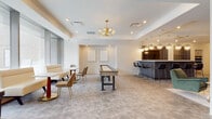 Located at 55 Public Square in the heart of Cleveland, Ohio, this single-floor amenity space offers a thoughtfully designed environment that blends comfort, functionality, and contemporary style. The layout encompasses several distinct zones that cater to a variety of social and recreational activities.

The lounge area is anchored by floor-to-ceiling windows that flood the space with natural light and frame striking views of the Cleveland cityscape. Leather sofas, plush green velvet armchairs, and intimate café-style seating arrangements create a welcoming atmosphere for relaxation and conversation. Gold-toned chandelier fixtures add a refined, modern elegance throughout.

Adjacent to the main lounge, a game and entertainment area features a full-length shuffleboard table as a centerpiece, complemented by café seating and a sleek bar counter clad in textured tile with pendant globe lighting overhead — ideal for social gatherings.

The fully equipped kitchen is a standout feature, outfitted with white shaker cabinetry, a blue subway tile backsplash, white quartz countertops, stainless steel appliances including double wall ovens, a full-size refrigerator, and a microwave. A large central island with coordinating blue tile detailing provides ample prep and gathering space beneath modern dome pendant lights.

Throughout, neutral toned flooring, recessed lighting, and a clean white palette create a cohesive, polished ambiance that is both inviting and functional.