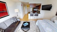 This well-appointed single-floor space offers a thoughtfully designed open-concept layout that seamlessly connects the living area and kitchen. The living room is furnished with a leather sofa, a plush area rug, and an ottoman, creating a comfortable and inviting atmosphere. A wall-mounted flat-screen TV adds a modern touch to the space.

The kitchen is a standout feature, showcasing rich dark wood cabinetry with a two-tone design — upper cabinets in warm wood tones complementing the darker lower units. Stainless steel appliances, including a refrigerator and microwave, provide a sleek, contemporary feel. A dark granite breakfast bar with modern bar stools serves as both a functional dining area and a visual divider between the kitchen and living space.

Light hardwood flooring flows throughout the open area, enhancing the bright and airy ambiance created by recessed lighting overhead. The entryway, visible from the living area, features multiple closet doors offering generous storage. Overall, the space is designed with clean lines, neutral tones, and modern finishes, making it a functional and stylish environment well-suited for both relaxing and entertaining.