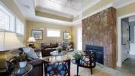 This inviting living space seamlessly combines rustic charm with contemporary comfort. The room's focal point is an impressive floor-to-ceiling stone fireplace, adding warmth and character. A tray ceiling with recessed lighting enhances the sense of space and sophistication. The seating arrangement includes a plush leather sofa and accent chairs, centered around a glass coffee table. Blue accents in the furniture and artwork provide a refreshing contrast to the neutral walls. Large windows allow natural light to flood the room, while framed landscape prints add a touch of nature-inspired decor. The space is further adorned with potted plants, bringing a hint of the outdoors inside. Hardwood flooring grounds the room, contributing to its overall cohesive and welcoming atmosphere. This thoughtfully designed living area strikes a perfect balance between comfort and style, creating an ideal setting for relaxation and entertaining.