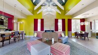 Located at 5109 Goldsboro Dr in Hampton, Virginia, this single-floor community clubhouse delivers a striking visual identity through its bold, energetic design. The space is anchored by a dramatic vaulted ceiling adorned with exposed white structural beams set against a deep teal backdrop, creating an impressive sense of vertical volume throughout the main lounge area. Vivid chartreuse-yellow walls paired with rich crimson curtains establish a lively, contemporary color palette that carries throughout the space.

The expansive main lounge is designed for comfort and social interaction, featuring modular striped seating arrangements, ottomans, and a central coffee table positioned around a sleek fireplace wall clad in neutral stone. Large windows dressed with two-toned red and tan drapes flood the interior with natural light and offer views of the surrounding neighborhood. Polished large-format tile flooring and patterned area rugs define distinct zones within the open layout.

A dedicated office area provides a functional workspace equipped with workstations, ergonomic seating, and a bold multicolored art installation on the yellow accent wall. Tall windows in this zone offer additional outdoor views. The entry area features soaring ceilings, chartreuse accent chairs, and glass-panel doors that invite natural light deep into the interior, completing a space that is both purposeful and visually dynamic.